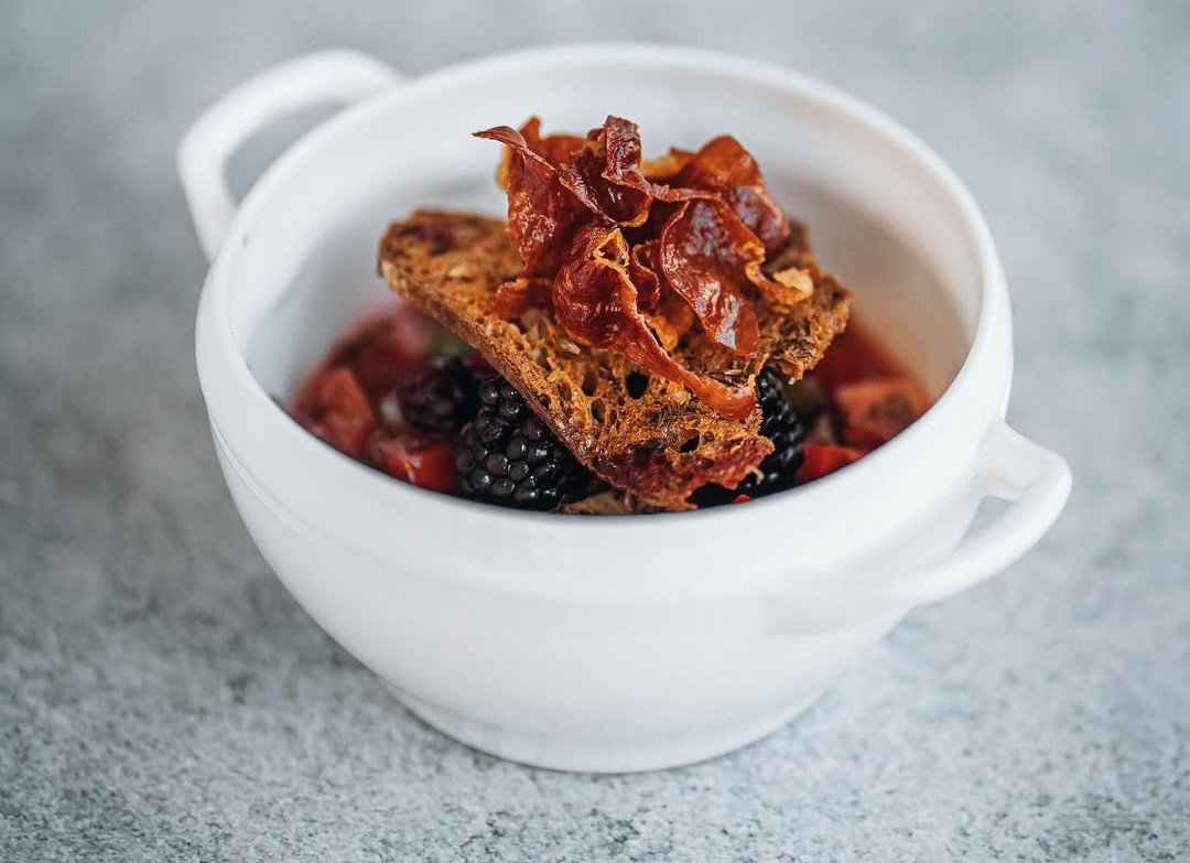 Tigela branca de barro com creme, torrada rústica, presunto e frutos vermelhos
