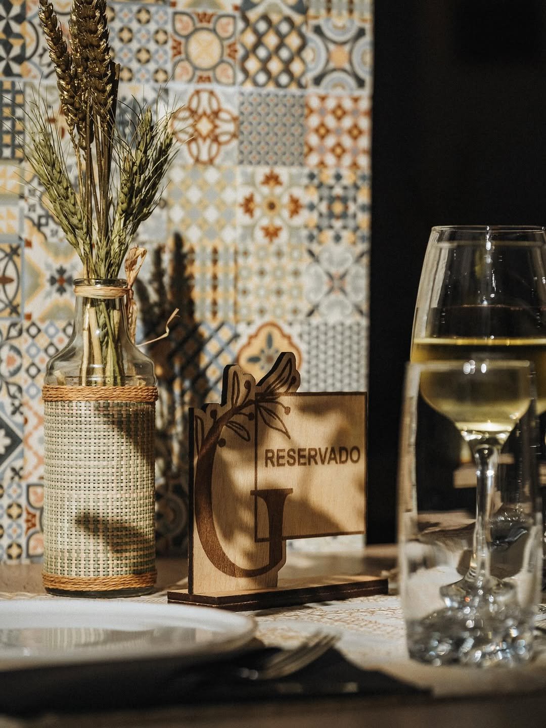 Mesa posta do restaurante com garrafa decorada, placa de madeira reservada e taças de vinho