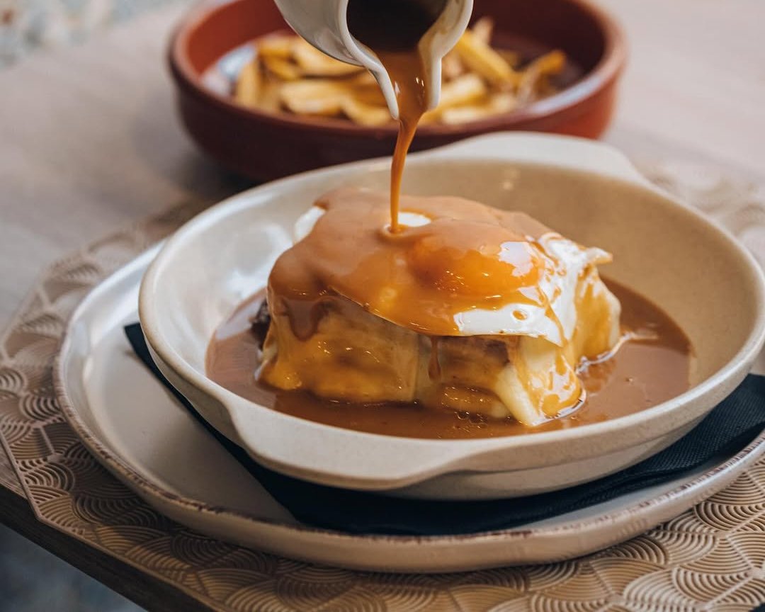 Francesinha com molho a ser servido sobre o prato, num prato de louça com batatas ao fundo